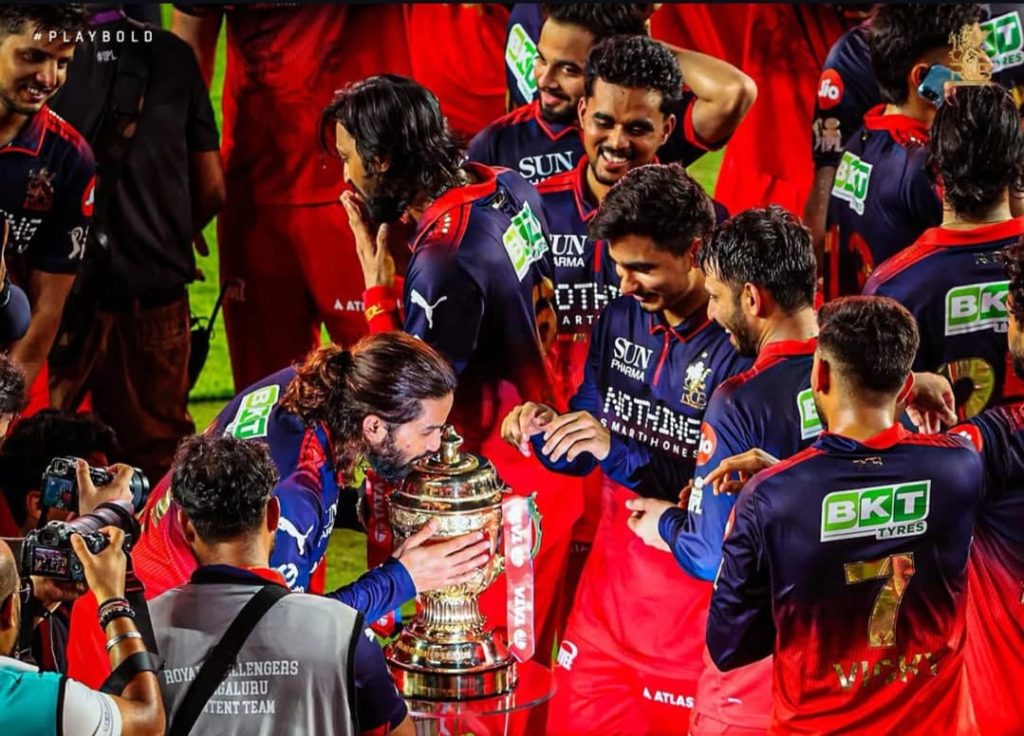 WhatsApp Image 2026-06-01 at 123935 AM - The Journalist News Team in blue and red uniforms surrounds a large trophy on the field, photographers capturing the moment.
