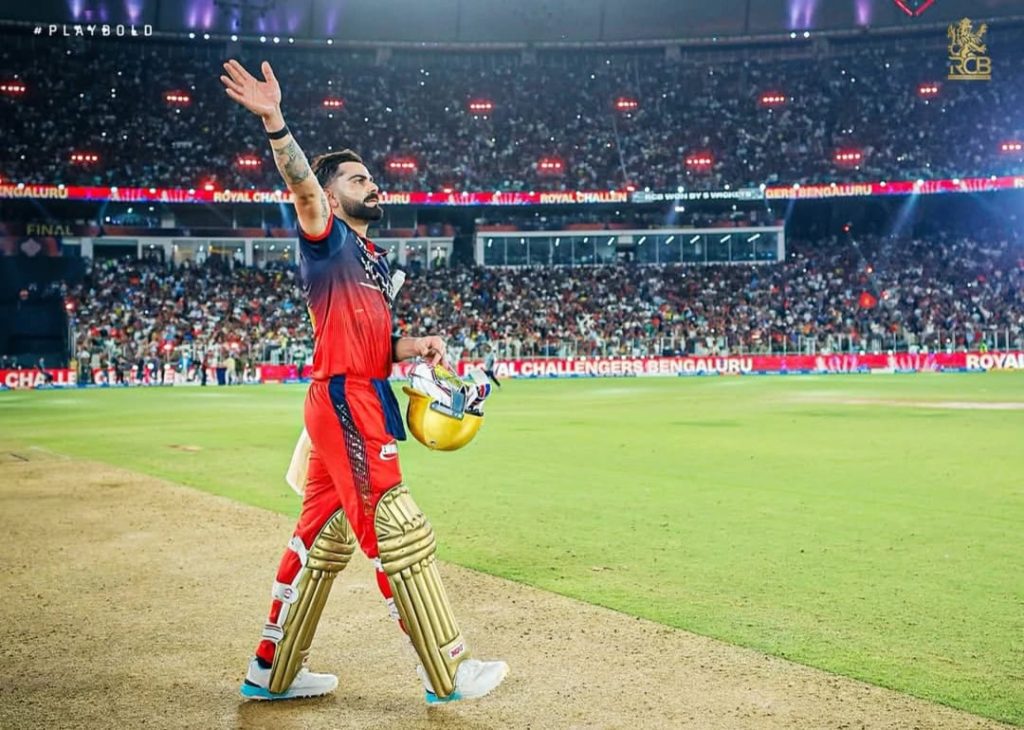 WhatsApp Image 2026-06-01 at 123936 AM - The Journalist News Cricketer in red-blue gear walks on the pitch, raises his right hand to the crowd, helmet in his left hand, gold pads gleaming as fans cheer.