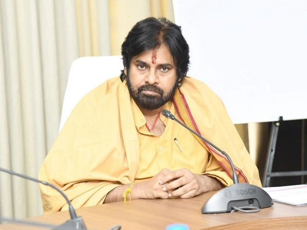 Bearded man in yellow robes sits at a conference table with a microphone in front, hands folded.