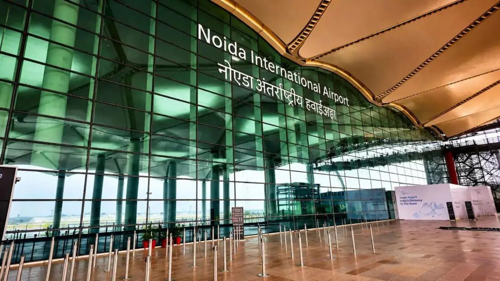 3e76231c2dca19b6698fb00d0b83ee84 - The Journalist News Front view of Noida International Airport with large green glass facade and a bold sign in English and Hindi reading 'Noida International Airport' and Hindi text beneath.