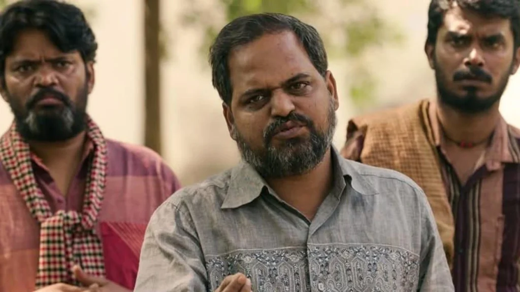 Three men stand outdoors with serious expressions, the central man speaking or addressing someone.