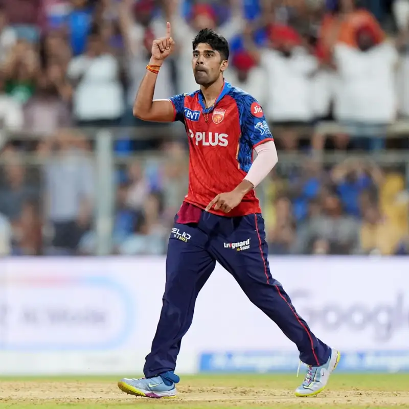 Cricketer in red and blue kit raises one finger in celebration on the pitch with a cheering crowd behind him