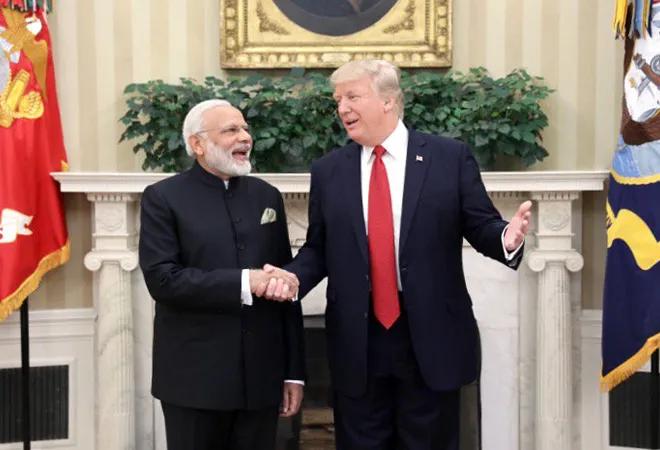 Two men in formal suits shake hands in a grand room with a fireplace and flags behind them, smiling at each other.