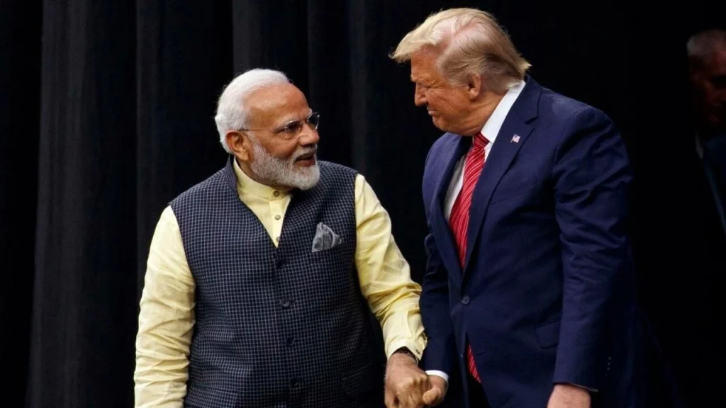 Two men in formal attire shake hands on a dark stage, smiling at each other.