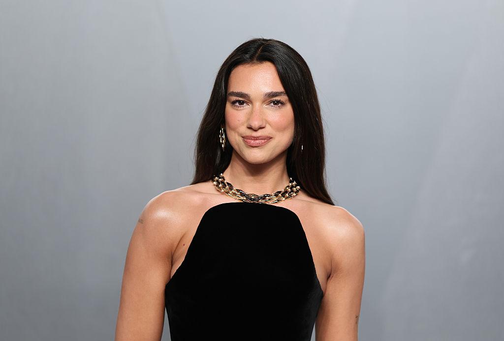 Woman with long dark hair wearing a black strapless dress and a gold chain necklace, posing against a gray backdrop.