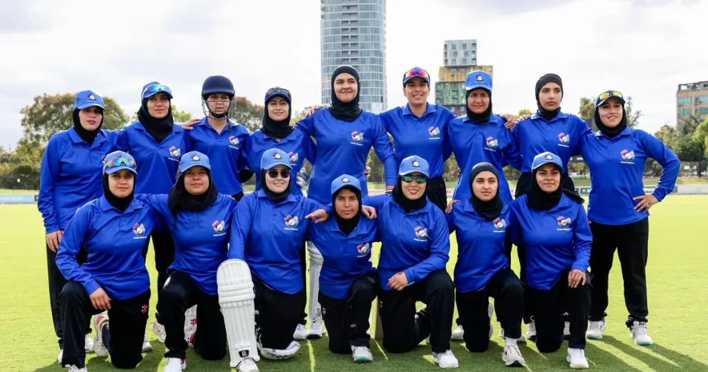 Group of Muslim women cricketers in matching blue uniforms posing on a sports field with city buildings in the background
