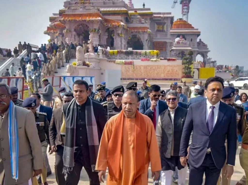 CM_Yogi_visit_Ayodhya__1781743050387_1781743050537_b3f43829-e5e2-4fbf-8e05-abe79449ae4a - The Journalist News Group of officials, including a man in orange robes, walking toward the camera at a temple construction site with crowds and security nearby.
