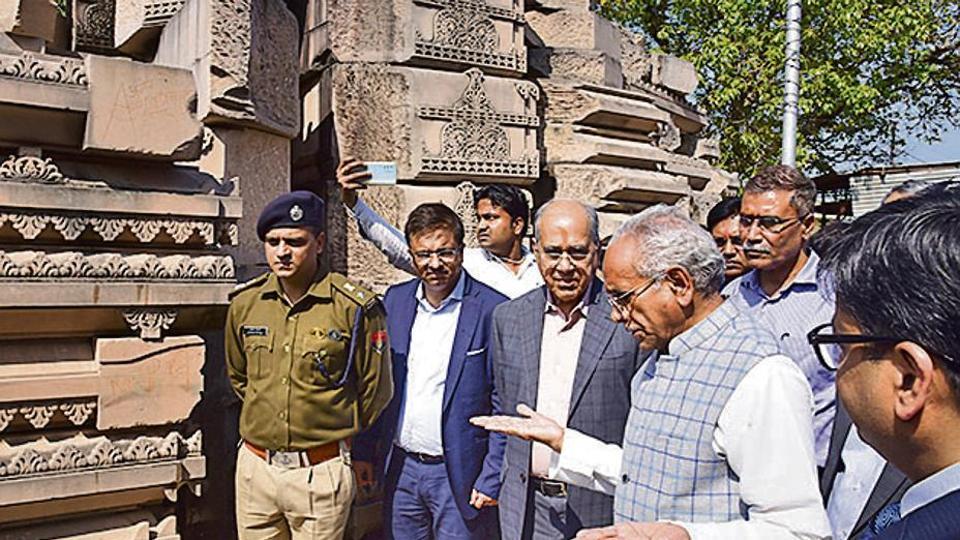 Group of officials and a police officer examine intricate stone carvings at an outdoor heritage site, one man gesturing.