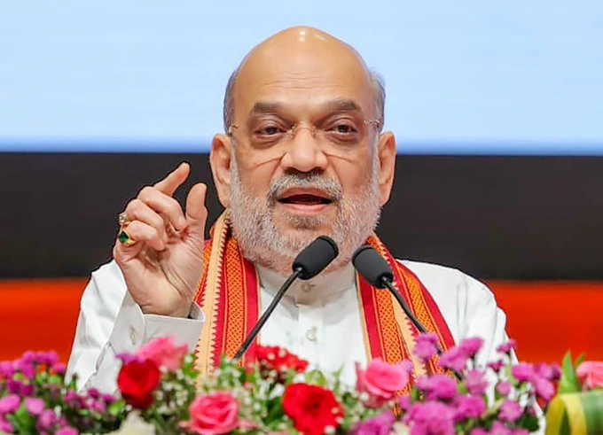 G1F6NzYW0AAZ3FK - The Journalist News Bald man with a white beard speaks into two microphones at a podium, wearing white traditional clothing and a red scarf, flowers in front.