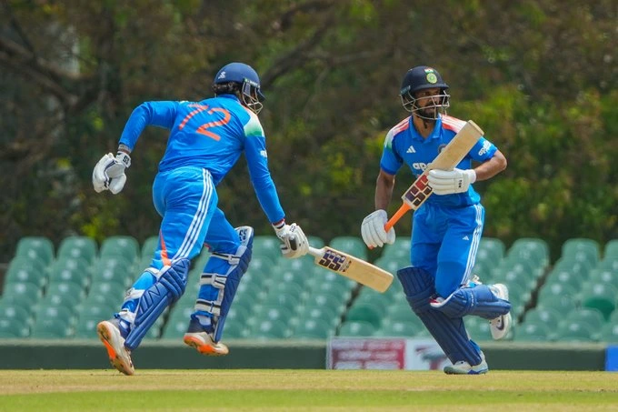 HK1Ug0eawAAYn3_ - The Journalist News Two cricket players in blue uniforms run between the wickets on a sunny field, wearing helmets and pads.
