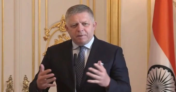 Man in a dark suit delivering a speech at a formal event, with an Indian flag and ornate gold decor in the background.