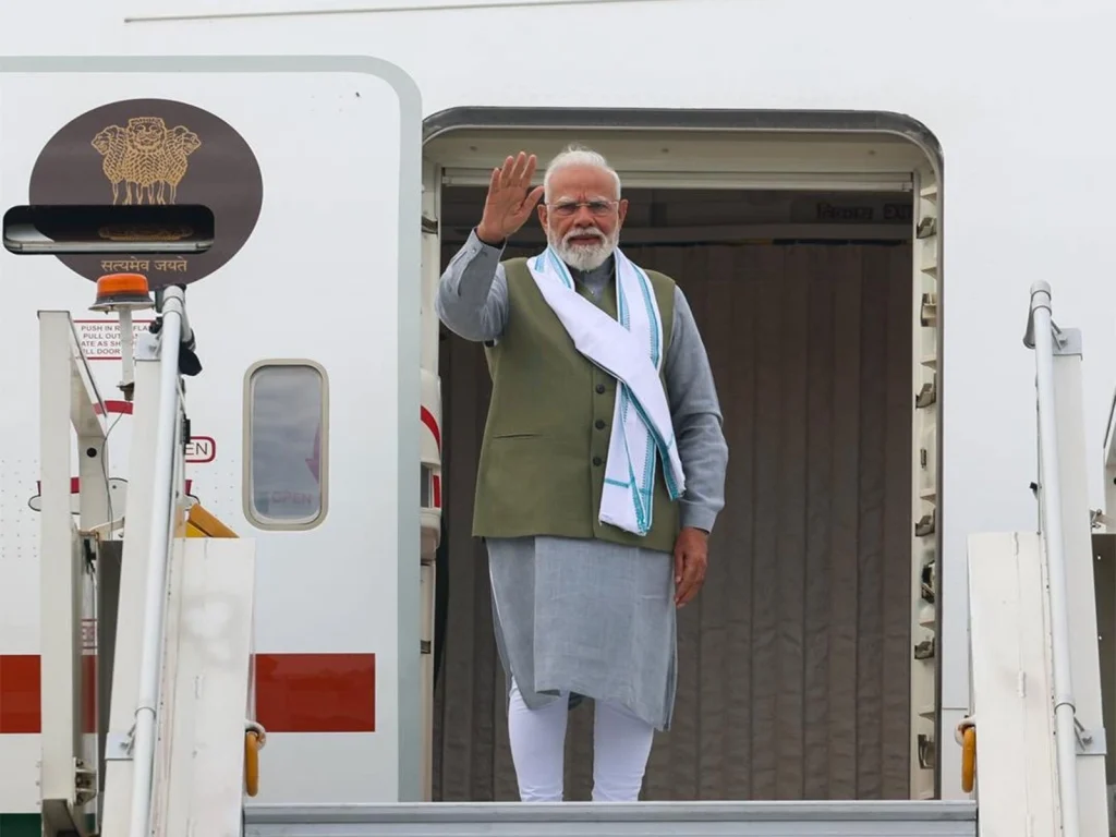Prime Minister Narendra Modi waves from the doorway of a white airplane, wearing a green vest and light gray kurta with a white scarf draped over his shoulder.