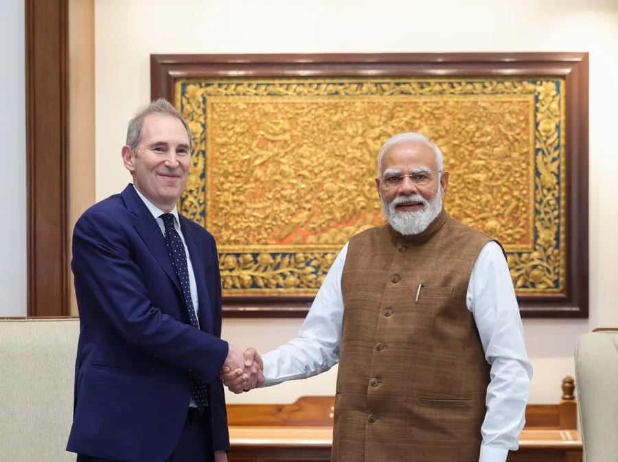 Two men in formal attire shake hands in a room with a large ornate gold-framed tapestry on the wall behind them, suggesting a formal meeting.