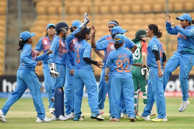 Indian women cricketers celebrate on the field, blue uniforms raised hands in a cheer.