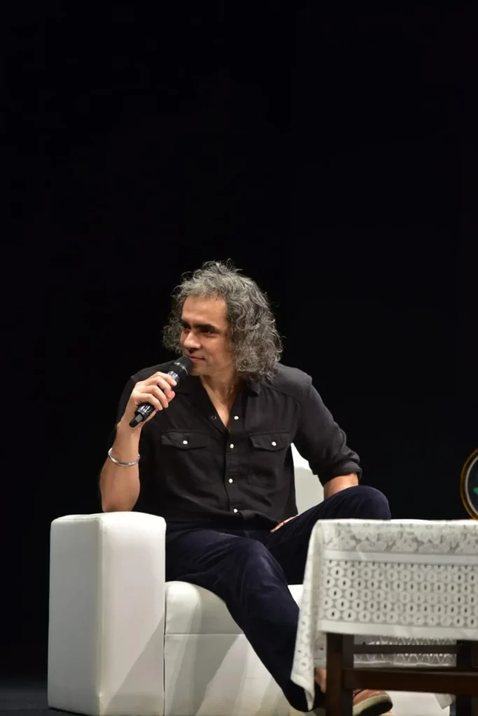 Man with gray, shoulder-length hair sits on a white armchair on a dark stage, speaking into a handheld microphone.