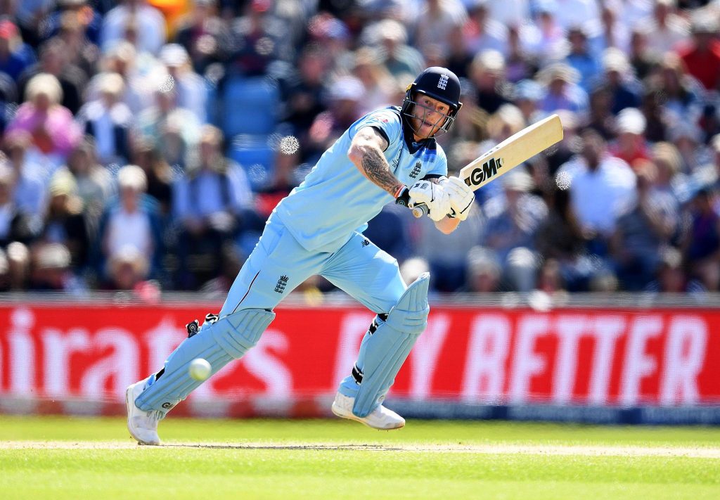 England cricketer in pale blue kit mid-swing, bat extended as ball approaches, crowd blurred in the background