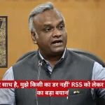 Man in a gray vest speaks at a microphone; Constitution of India book appears on the left.