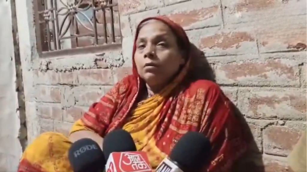 Woman in a red and yellow sari sits against a brick wall, speaking to reporters holding microphones in front of her.
