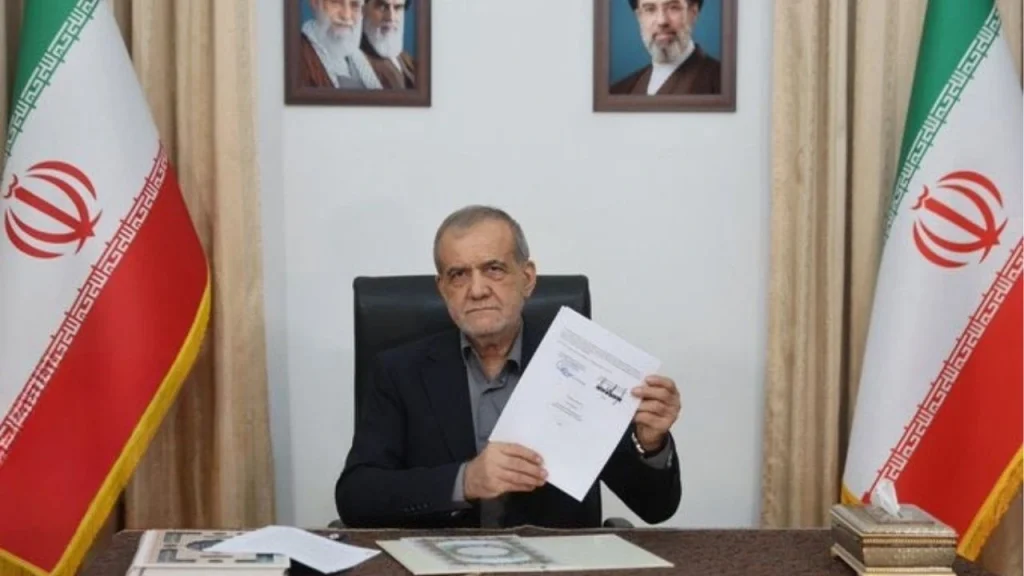 Older man in a suit sits at a desk, holding a document toward the camera with two Iranian flags in the background.