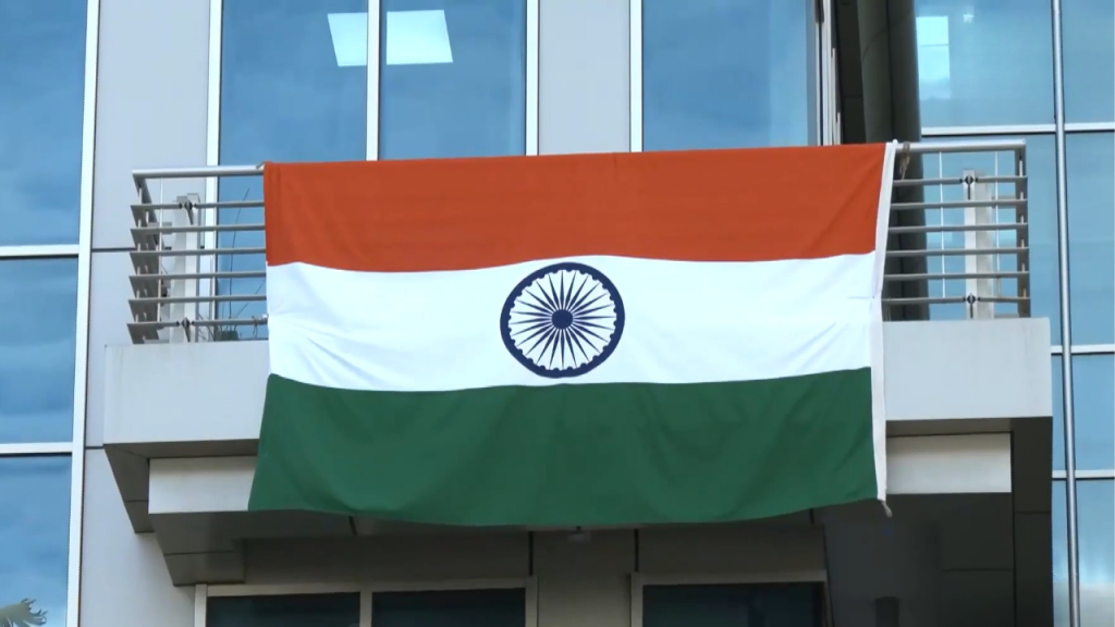 Indian flag draped over a balcony railing on a modern glass building, displaying saffron, white, green stripes with the Ashoka Chakra in the center.