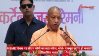 CM Yogi Adityanath delivering a speech at a MSME Day event, with two microphones and a security guard behind, backdrop with Hindi text.