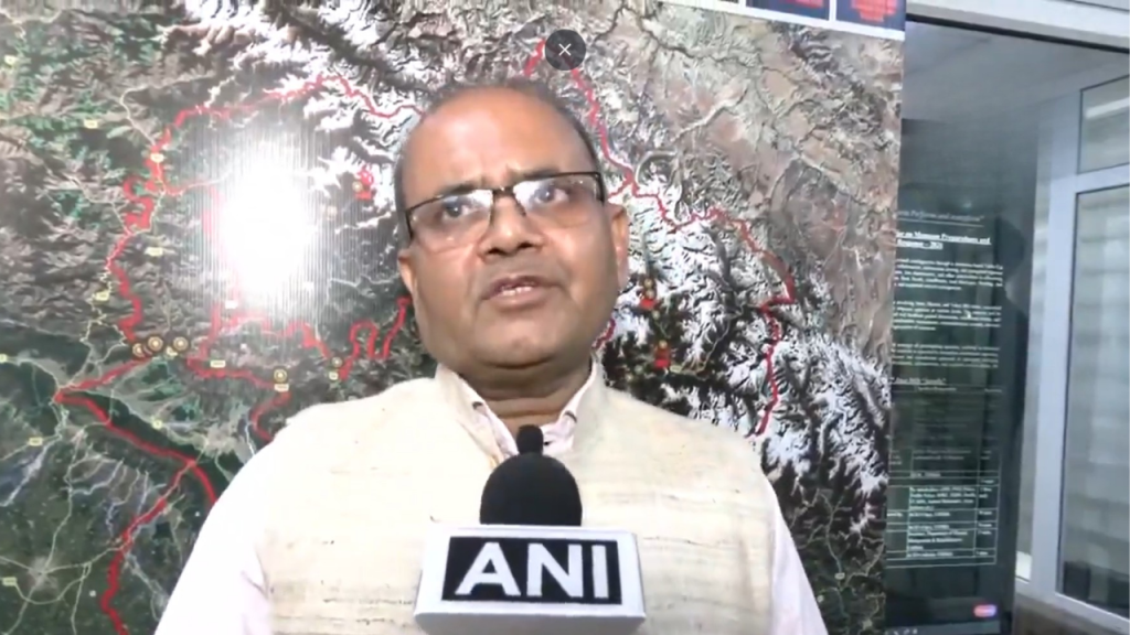 Indian man wearing a beige jacket speaks into an ANI microphone during a street/interview setting with a large map backdrop behind him.