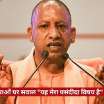 Speaker at a podium addressing a press conference, with two microphones and a red Hindi news banner about land mafia questions at the bottom.