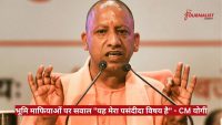 Speaker at a podium addressing a press conference, with two microphones and a red Hindi news banner about land mafia questions at the bottom.