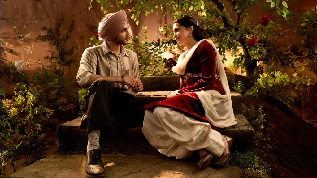 A man in a turban and a woman in a red and white traditional dress sit on stone steps, smiling at each other in a lush garden.