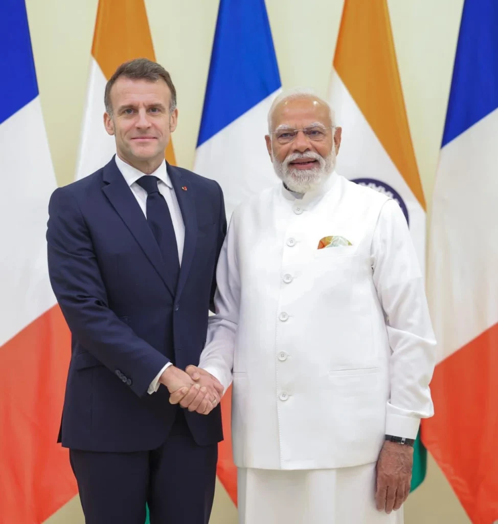 Two men in formal attire shake hands in front of colorful flags, signaling a diplomatic meeting.