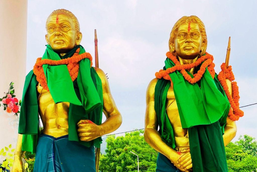 Two gilded statues of standing figures wearing green cloth and orange garlands, holding spears, outdoors with trees in the background.