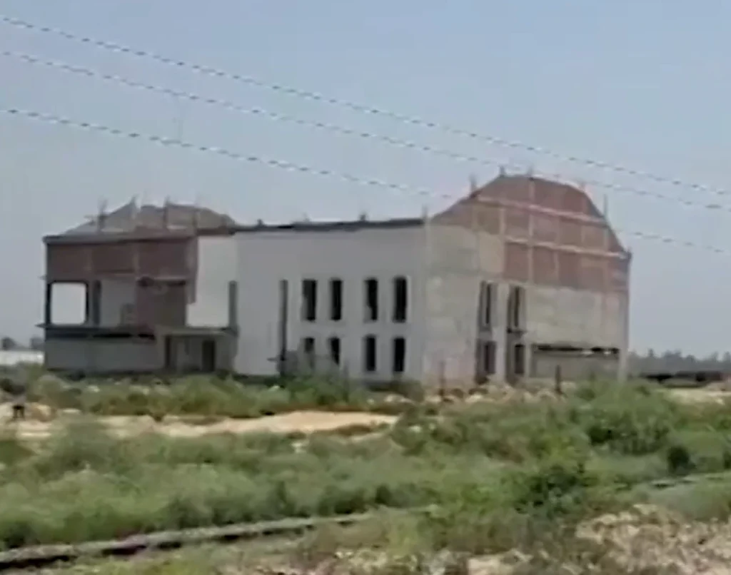 Unfinished two-story building under construction in an open field, with barbed-wire fencing in the background.