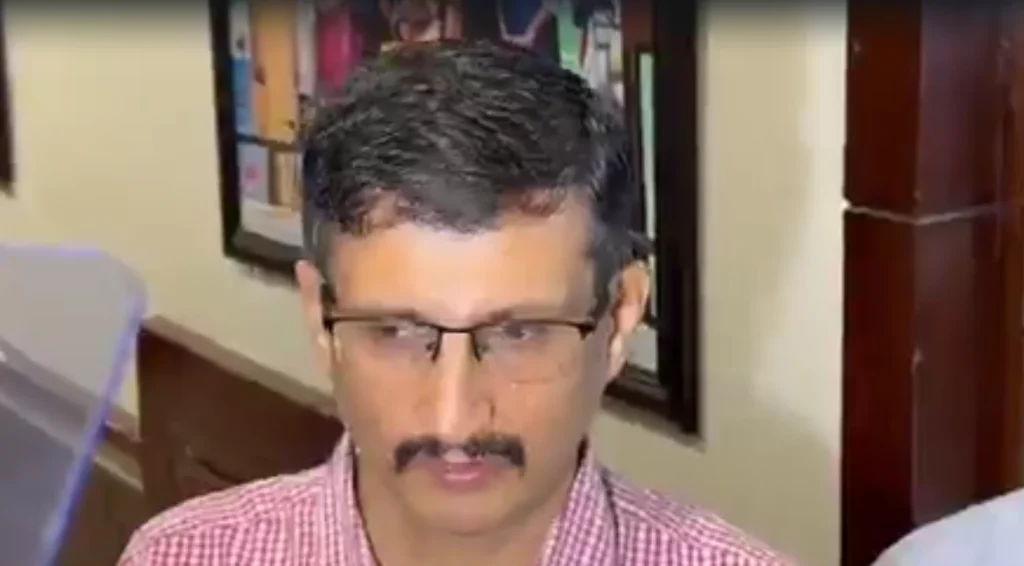 Screenshot 2026-06-23 145256 - The Journalist News Man with dark hair, glasses, and a mustache wearing a red checkered shirt indoors (near a wall with a framed picture).