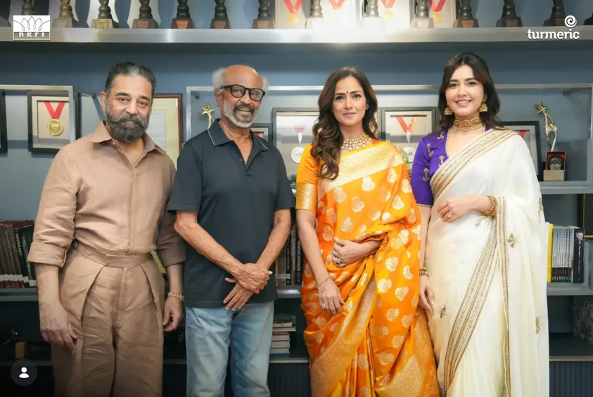 Four people posing in front of a trophy-filled shelf; two men flank two women in colorful sarees, smiling at the camera.