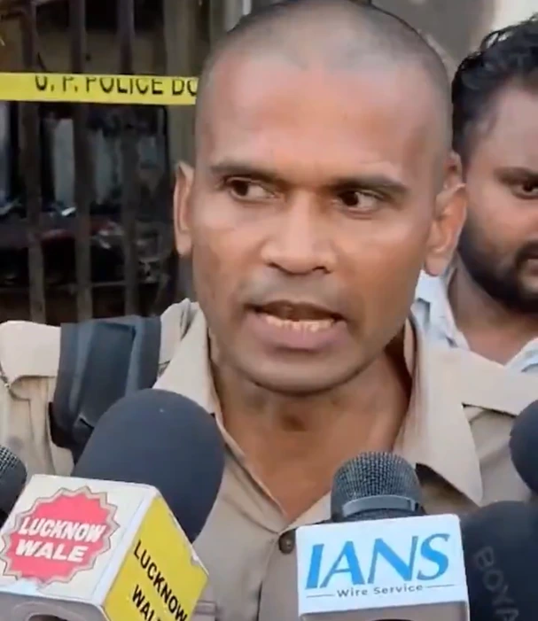 Police officer in a tan uniform speaks to reporters, microphones in front labeled IANS and Lucknow Wale, with yellow police tape in the background.