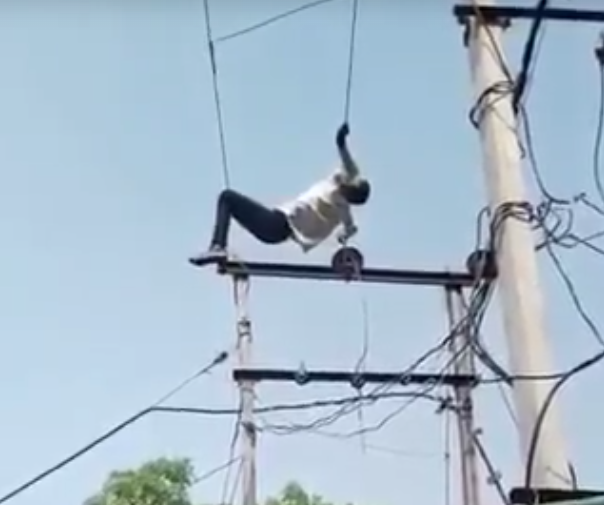 Person in a white shirt and dark pants balances on a high slackline between utility poles, gripping the rope with one hand against a clear blue sky to the side of the wires.] Getter user intent: informative and concise