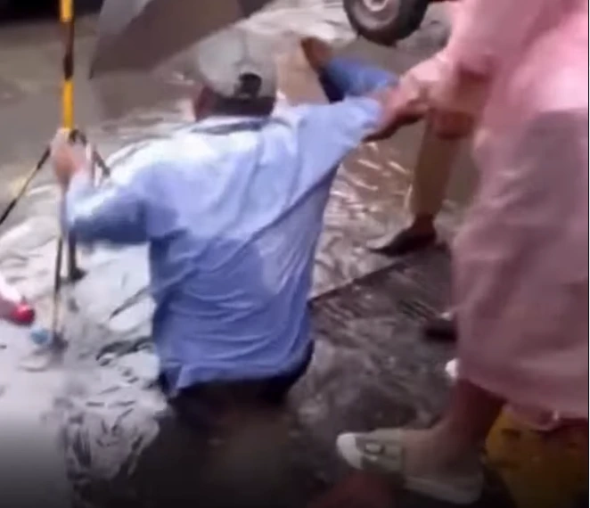 Man in a blue shirt kneels in knee-deep floodwater while others reach out to pull him to safety.