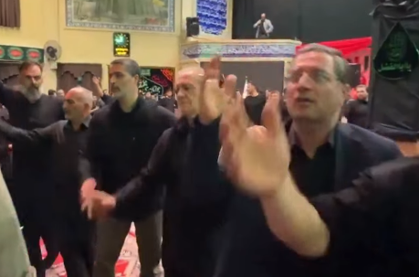 Several men in black shirts stand shoulder to shoulder, raising their hands in a coordinated group gesture inside a hall with ornate decor and a stage in the background.