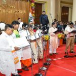 Group of people in traditional Indian attire singing into microphones on a red-stage during a formal ceremony, with an Indian flag and speakers in the background.