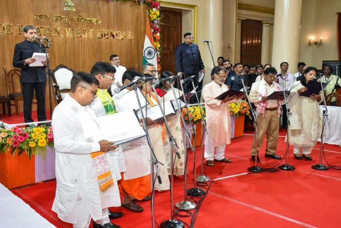 Group of people in traditional Indian attire singing into microphones on a red-stage during a formal ceremony, with an Indian flag and speakers in the background.