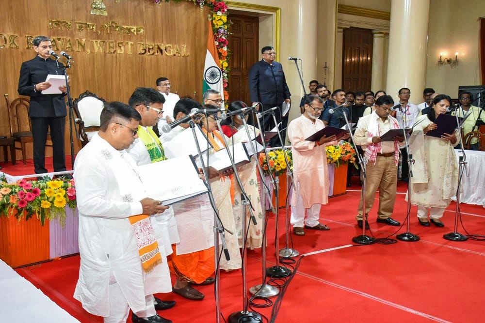 Group of people in traditional Indian attire singing into microphones on a red-stage during a formal ceremony, with an Indian flag and speakers in the background.