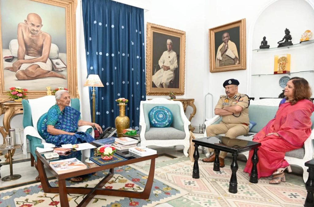 WhatsApp Image 2026-06-02 at 111953 PM - The Journalist News Elderly woman in a blue sari sits in a teal chair across from a police officer in uniform and a woman in a pink sari in a richly decorated living room with large portraits on the wall.