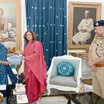 Two women exchange a booklet in a formal room as a police officer stands nearby, with large portraits on the walls behind them.