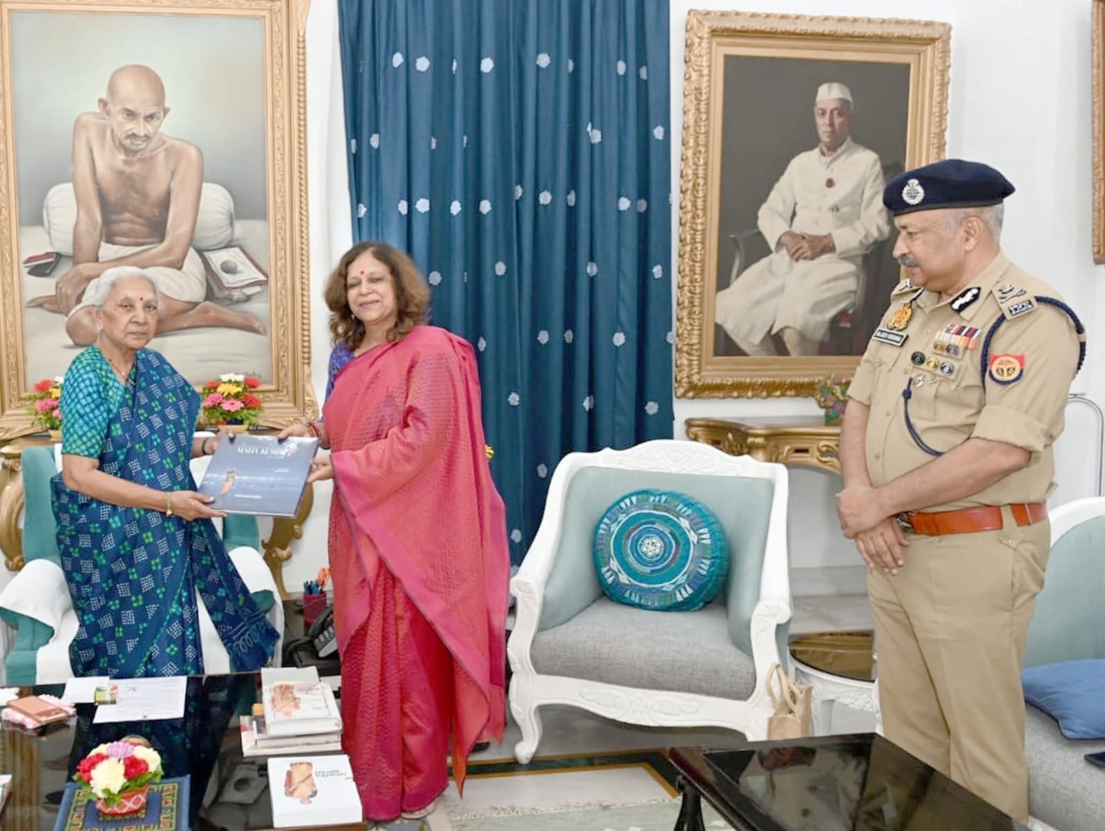 Two women exchange a booklet in a formal room as a police officer stands nearby, with large portraits on the walls behind them.