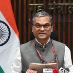 Indian government official speaking at a press conference, with flag and microphones in front.