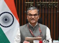 Indian government official speaking at a press conference, with flag and microphones in front.