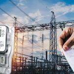 Electric meter in the foreground with a power substation and a hand holding a receipt in the background, suggesting energy billing or usage.