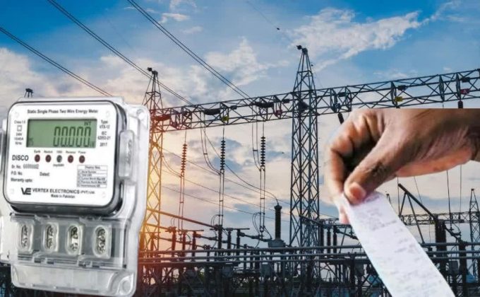 Electric meter in the foreground with a power substation and a hand holding a receipt in the background, suggesting energy billing or usage.