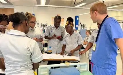 Diverse group of medical students in white uniforms gathered around a patient bed in a clinical skills room, examining a chart and discussing care.