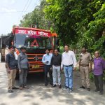 Seven men stand beside an orange transport truck on a sunny road, with lush green trees to the right; the truck has Hindi text on the windshield (likely a transport department name).